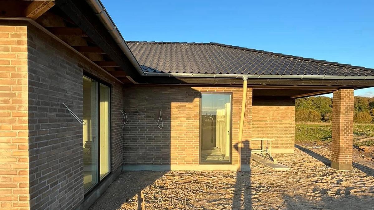 Nybygget hus med mursten, store vinduer og tag i sort, beliggende i et åbent landskab under blå himmel.