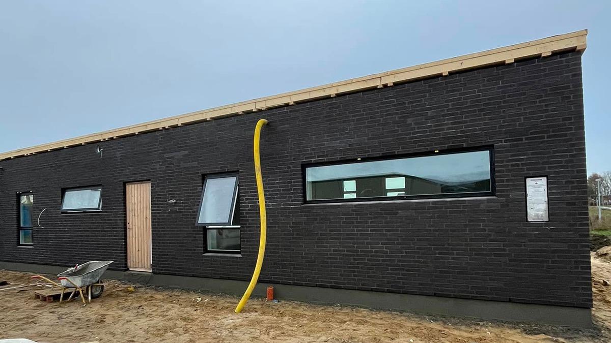 Moderne sort murstenshus med store vinduer og en trædør, under konstruktion med byggeudstyr i forgrunden.