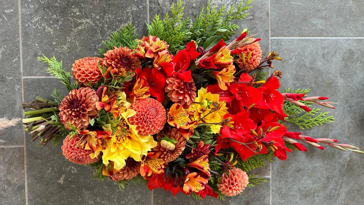 En farverig buket med orange, røde og gule blomster samt grønne blade på en grå fliseoverflade.