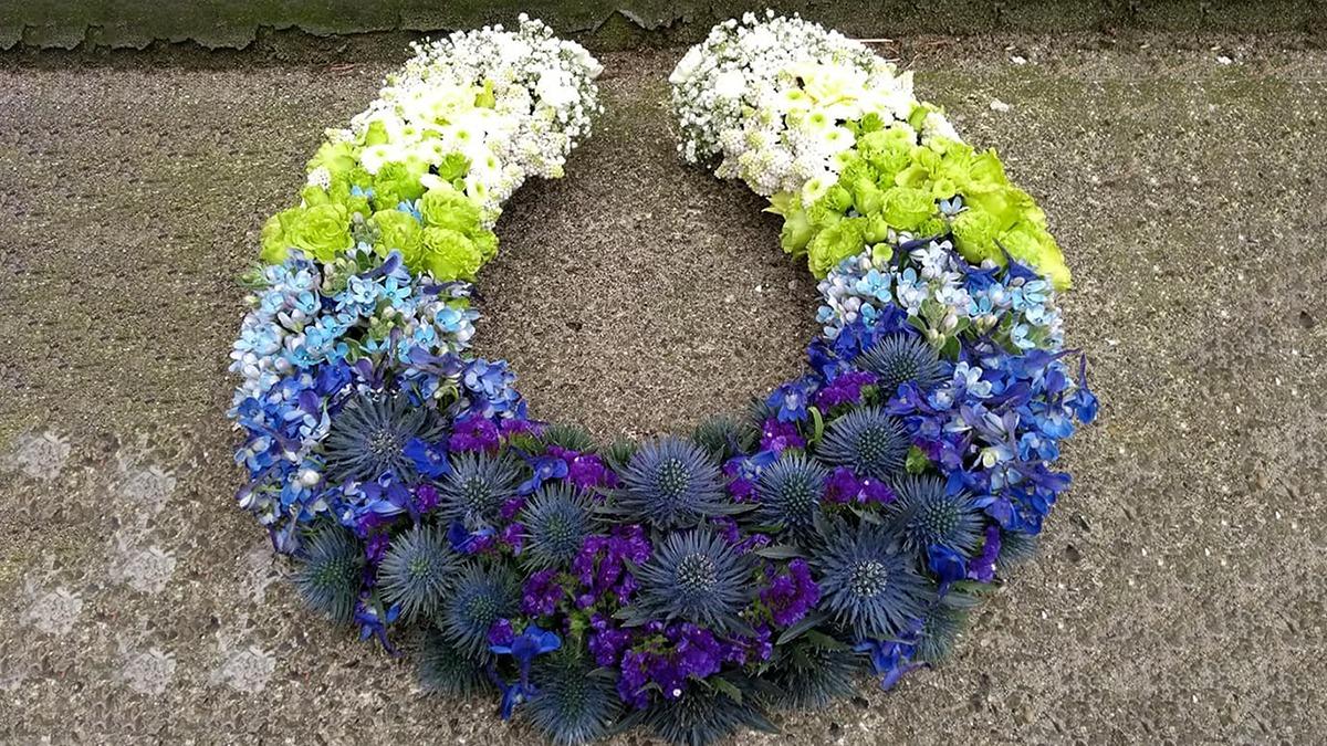 En farverig blomsterkrans i nuancer af blå, grøn og hvid, arrangeret i en halvmåneform på en grå overflade.