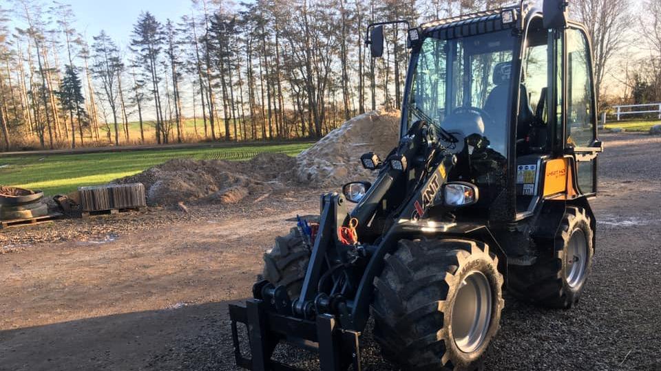 En sort minilæsser parkeret på en grusvej med træer i baggrunden og en bunke grus ved siden af.