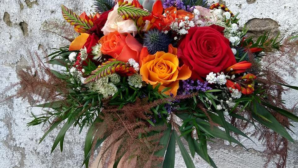 En farverig buket med røde, orange og lyserøde roser samt forskellige grønne planter og blomster.