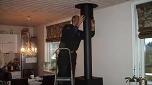 En mand står på en stige og installerer en sort skorsten i et køkken med vinduer og planter.