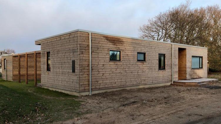 Moderne træhus med store vinduer og en indgang, omgivet af græs og træer.