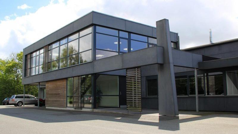 Moderne bygning med store vinduer og en træfacade, beliggende på en grusvej.
