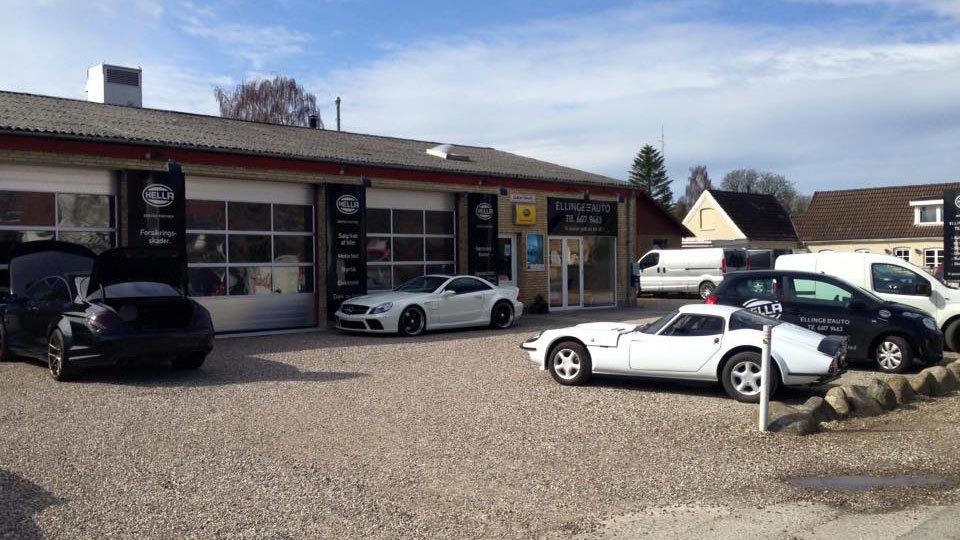 Bilforhandler med flere biler parkeret foran bygningen, herunder sportsvogne og varevogne.