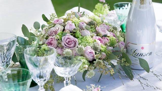En smuk buket med lyserøde roser og grønne planter på et festligt bord med glas og champagne.