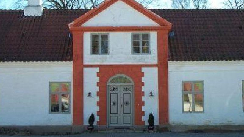 En hvid bygning med rød facade og to døre, omgivet af træer og en klar himmel.