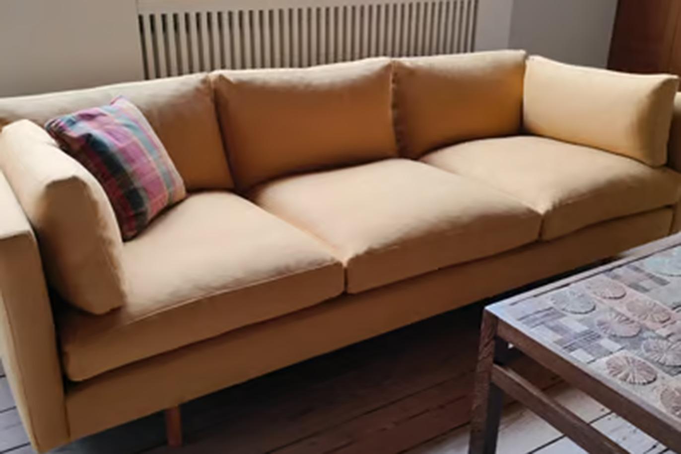 En gul sofa med bløde puder og et farverigt tæppe på armlænet, placeret ved siden af et træbord.