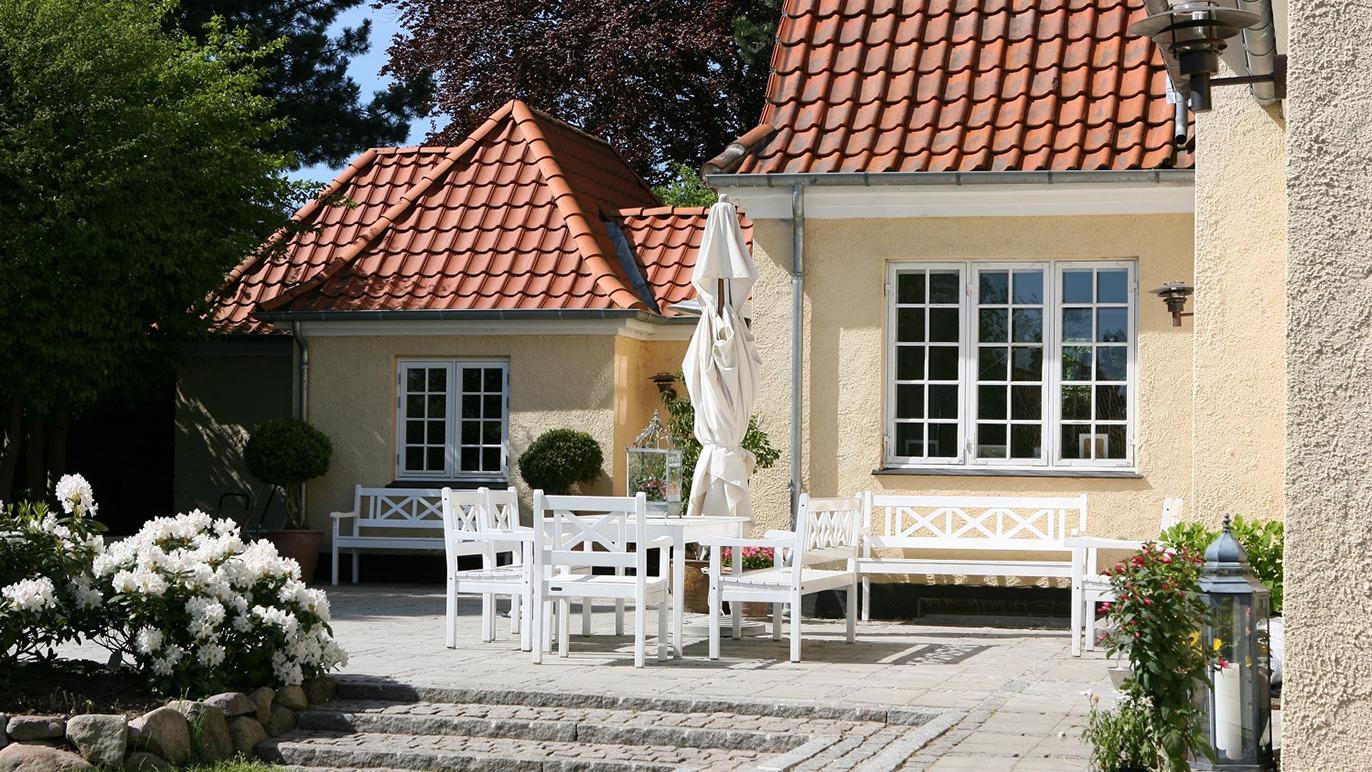 Et hyggeligt hus med rød tagsten, hvidt havemøblement og blomster i haven på en solrig dag.