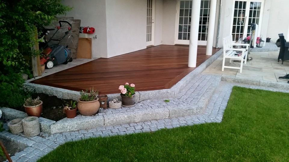 Terrasse med trædæk, blomsterkrukker og græsplæne ved en husvæg.