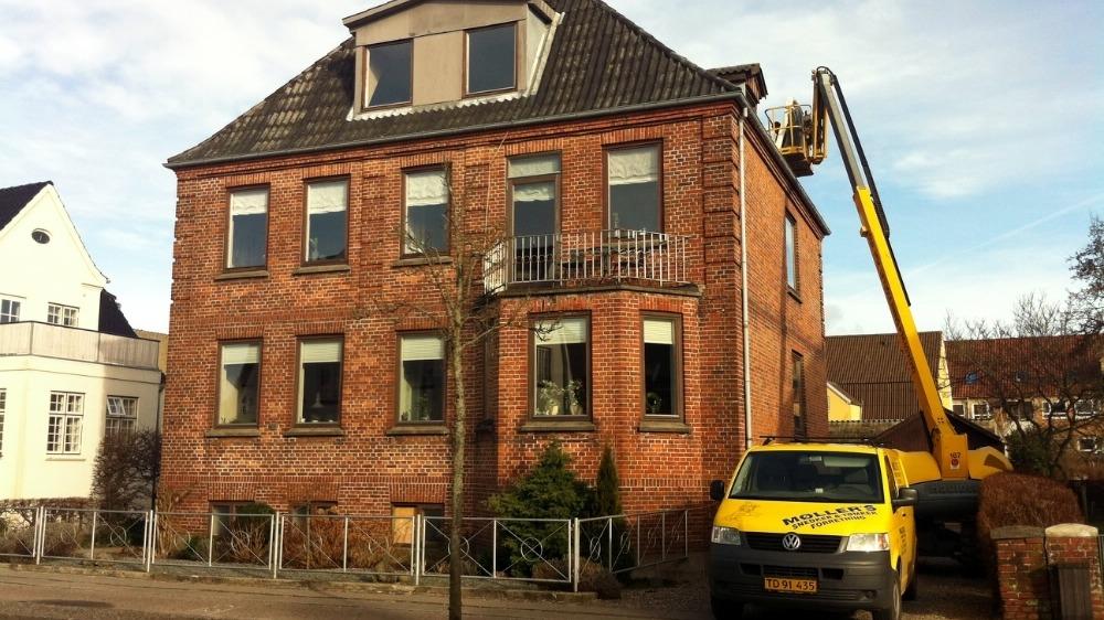 En murstensbygning med mange vinduer og en gul varevogn parkeret foran, mens en lift arbejder på taget.
