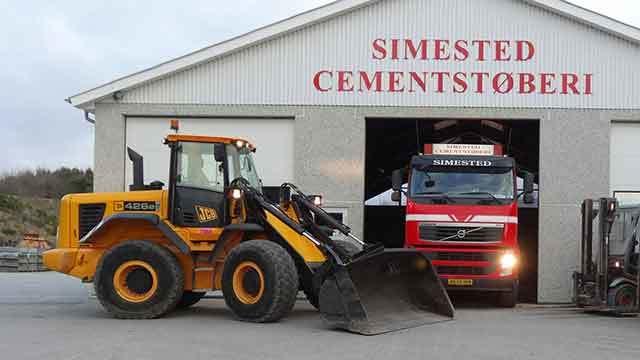 En gule gravemaskine står foran en cementfabrik med en rød lastbil i indkørslen.