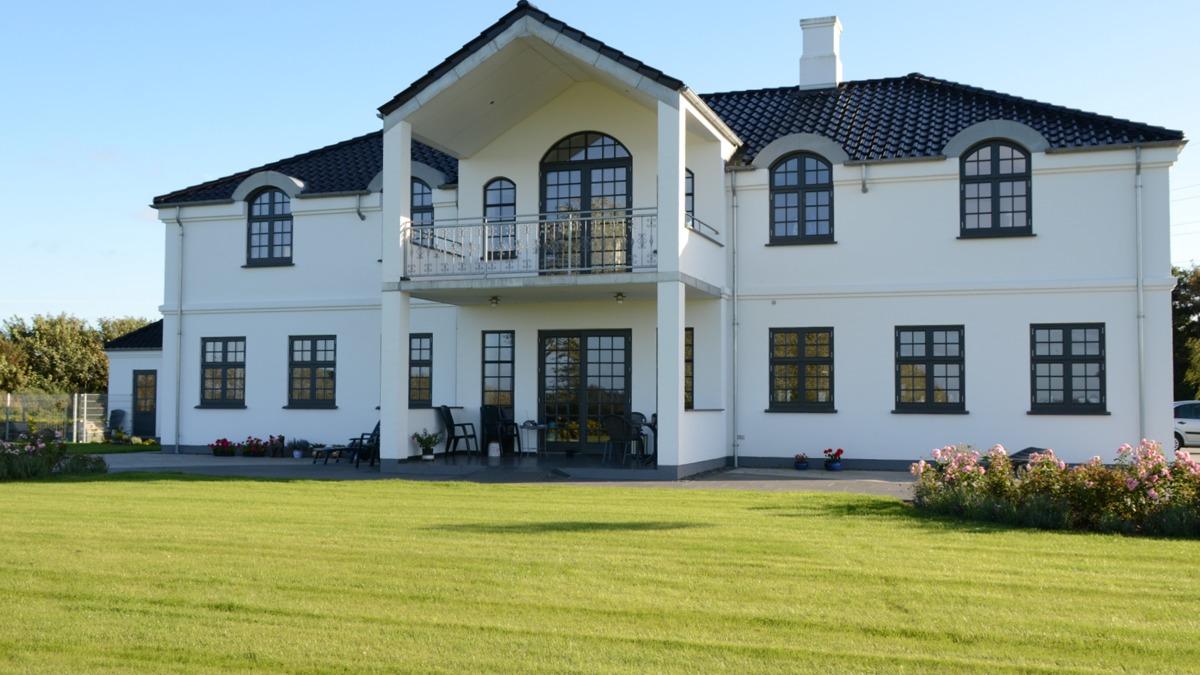 Et stort, moderne hus med hvid facade, sorte vinduer og en balkon, omgivet af en velplejet græsplæne.