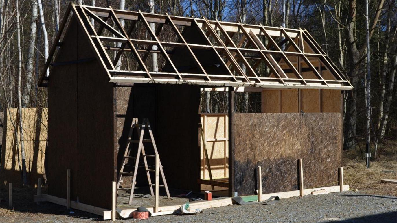 Et ufuldstændigt hus med tagkonstruktion, der står på en grusvej omgivet af træer.