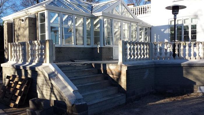 Glasveranda med trapper og balustre, omgivet af en stor terrasse i sollys.