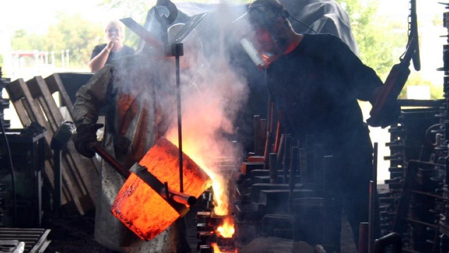 To mænd arbejder i et smedje, hvor de hælder smeltet metal fra en gryde i en form. Røg og flammer er synlige.