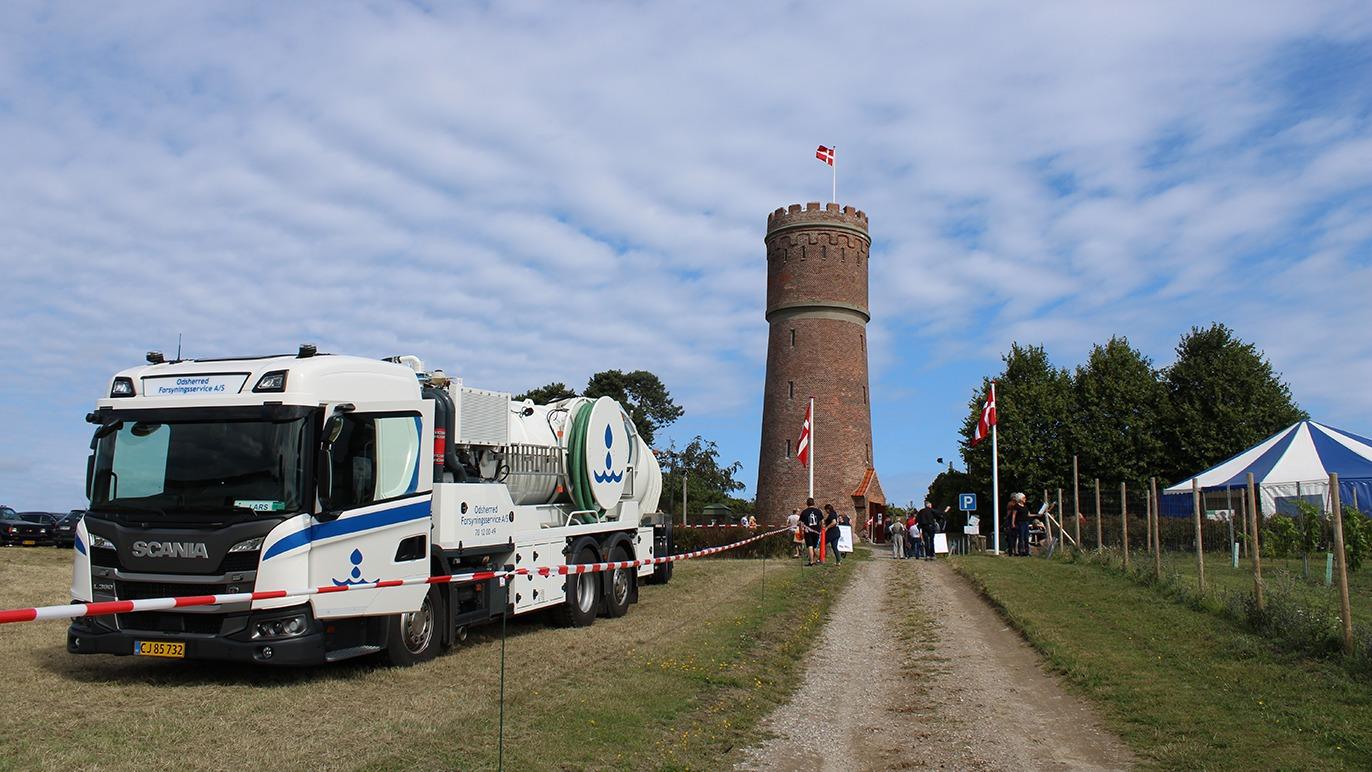 En lastbil med kran står ved en rund bygning, mens folk går mod tårnet under en blå himmel.