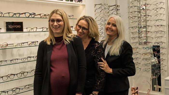 Tre kvinder står sammen foran en væg med briller i en optikerbutik. Den gravide kvinde smiler.
