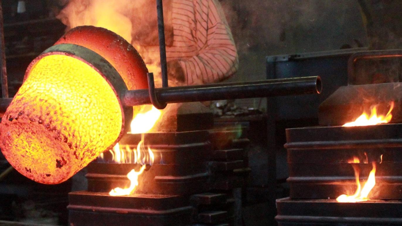 En smed, der hælder glødende metal fra en gryde i forme, omgivet af flammer i et smedeværksted.