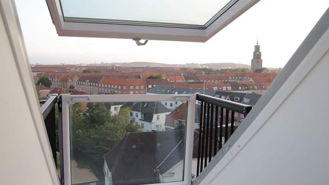 Udsigt fra en tagterrasse med åbent vindue, der viser byens tage og en kirketårn i baggrunden.