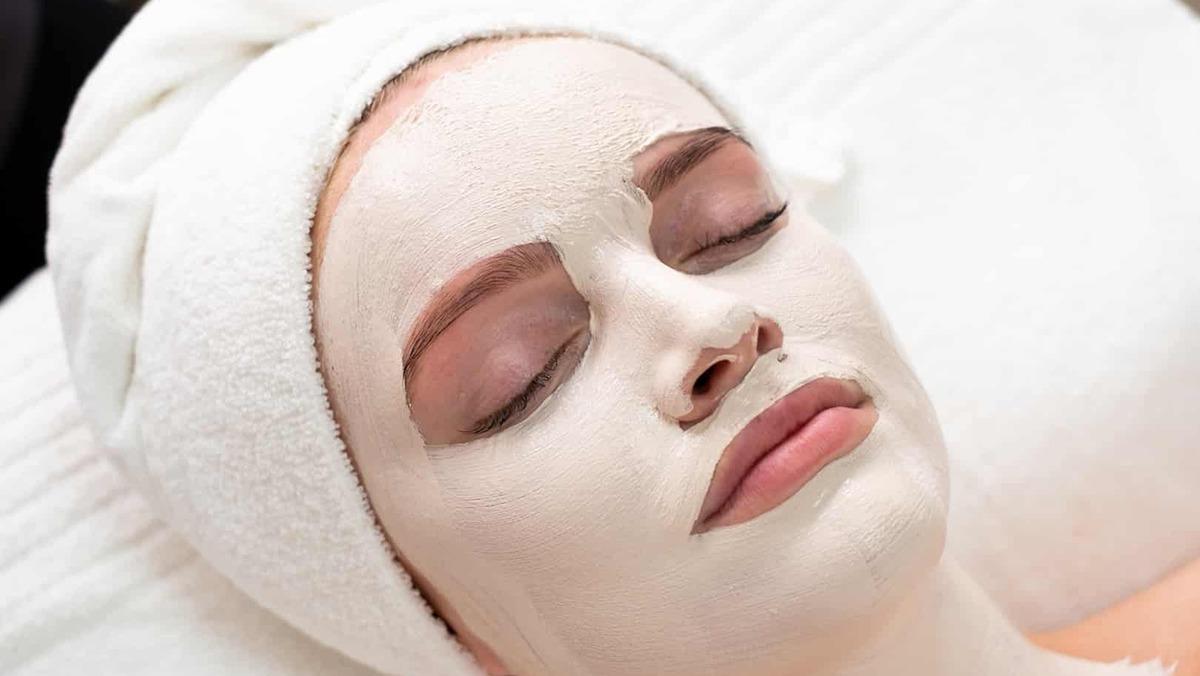 Kvinde med ansigtsmaske ligger afslappet på en briks i et spa, iført hvidt håndklæde om håret.
