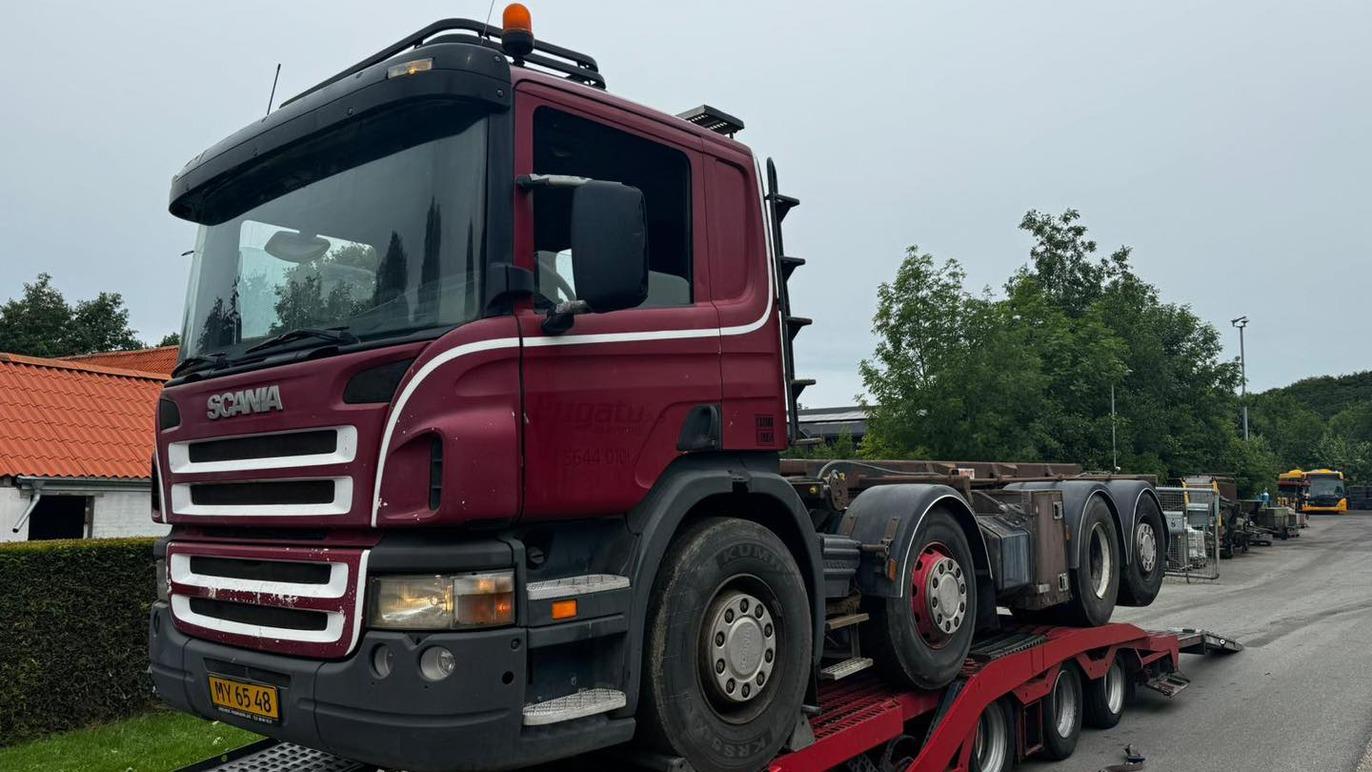 En rød Scania lastbil på en trailer, parkeret ved en vej med træer og bygninger i baggrunden.