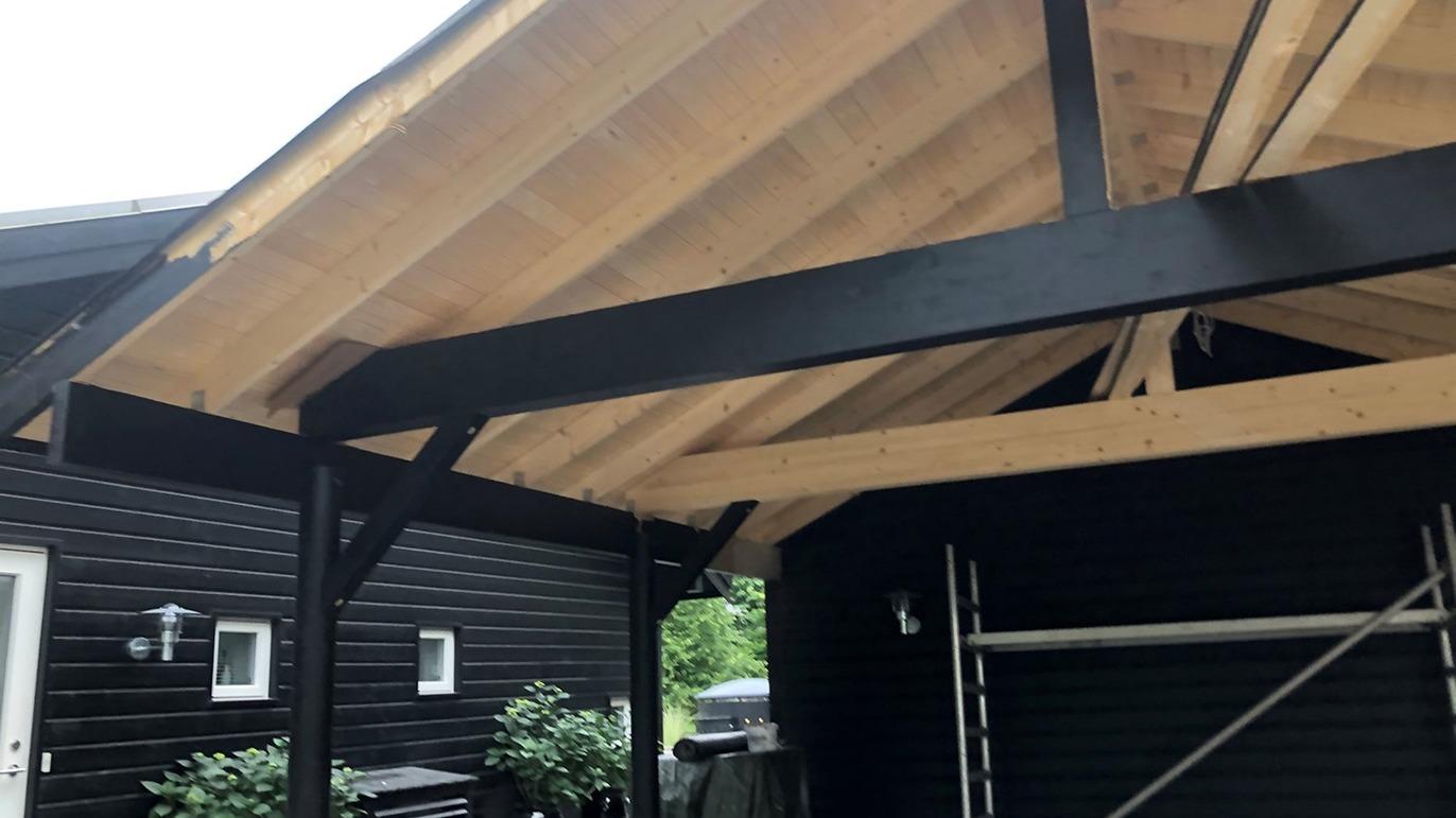 Tagkonstruktion med synlige træbjælker under en carport i sortmalet hus.