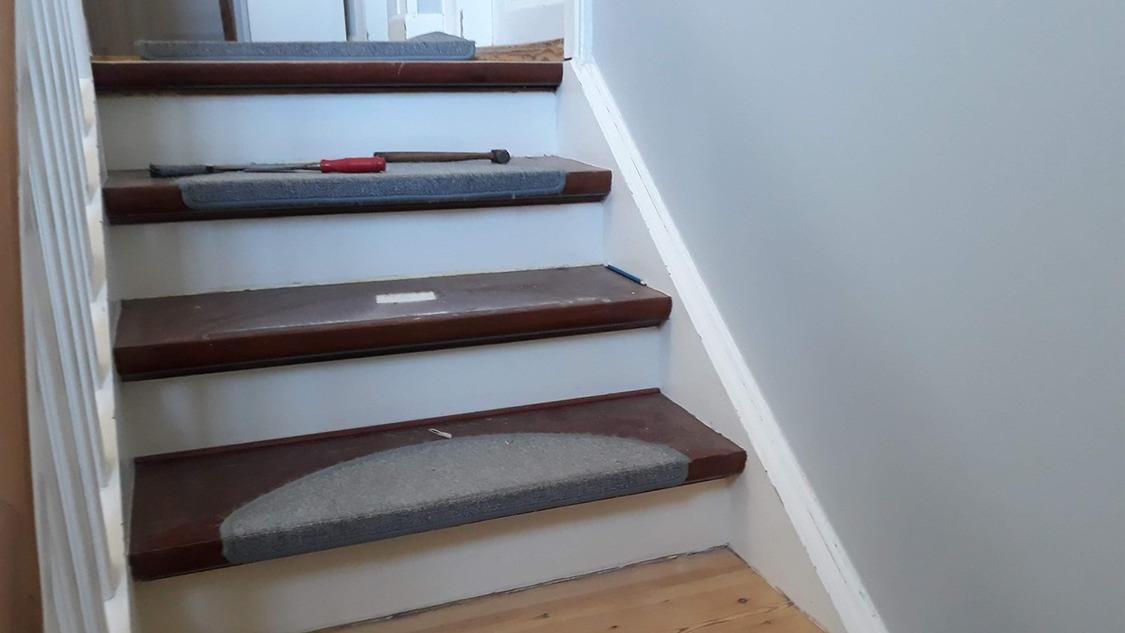 Trappe med grå tæpper på trin og en rød genstand på det øverste trin. Væggen er lys og trappen er i træ.