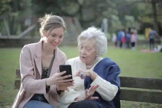 To kvinder, en ung og en ældre, sidder på en bænk i parken og ser på en smartphone sammen.