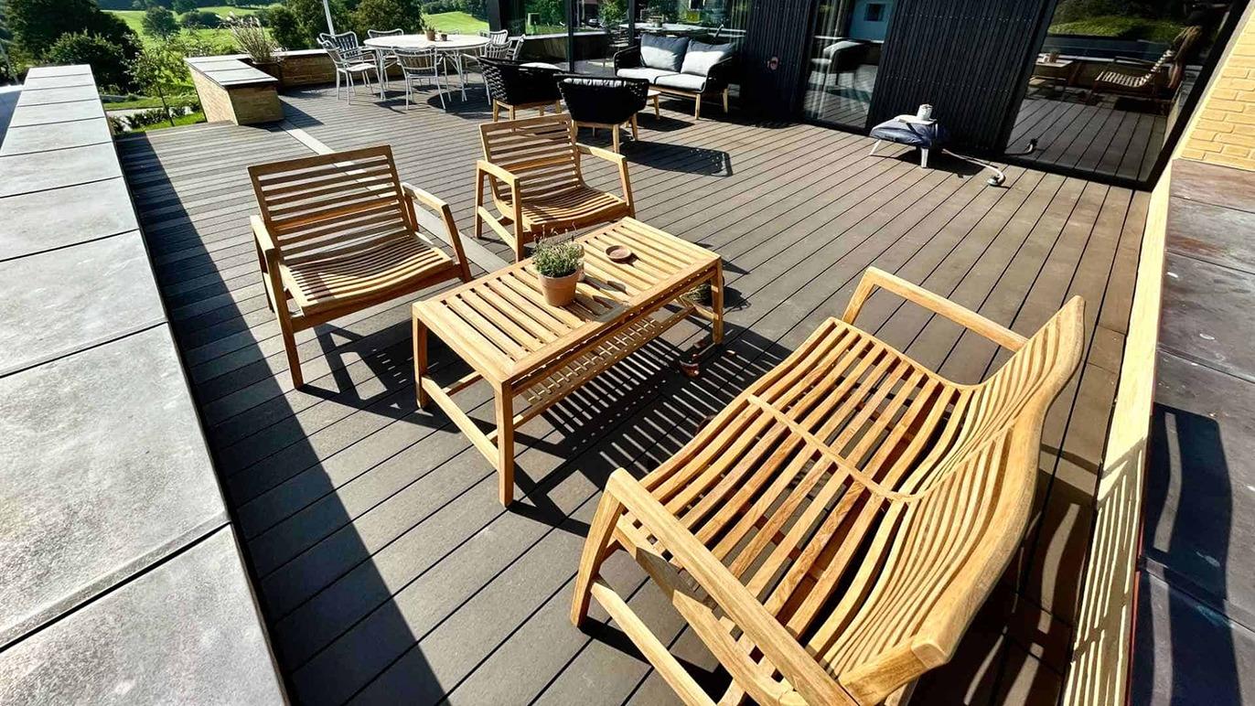 En solrig terrasse med træmøbler, herunder to stole og et bord, omgivet af grønne områder.