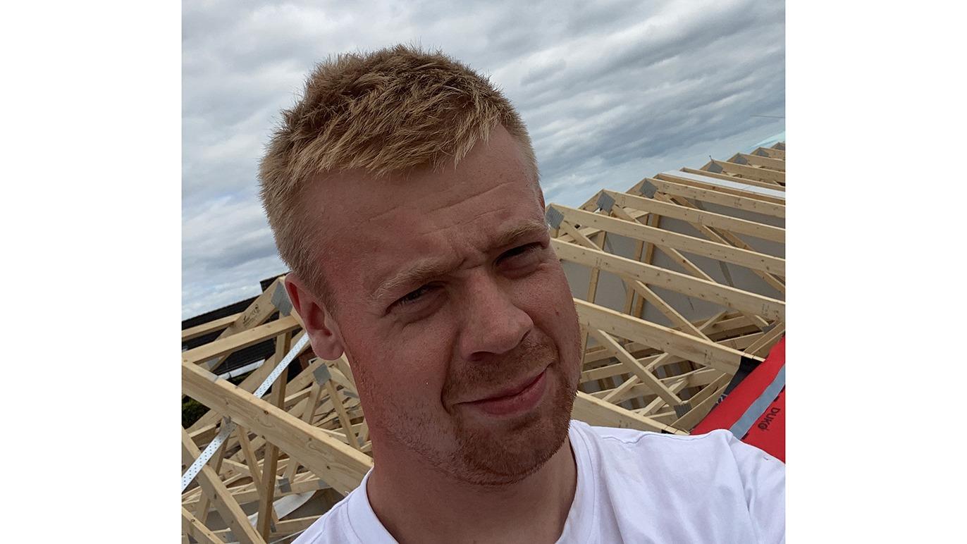 En ung mand står på en byggeplads med trærammer i baggrunden under en overskyet himmel.