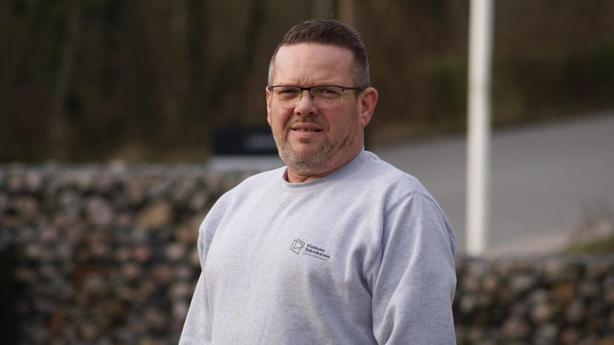 En mand i grå sweatshirt står udendørs med en stenmur i baggrunden.