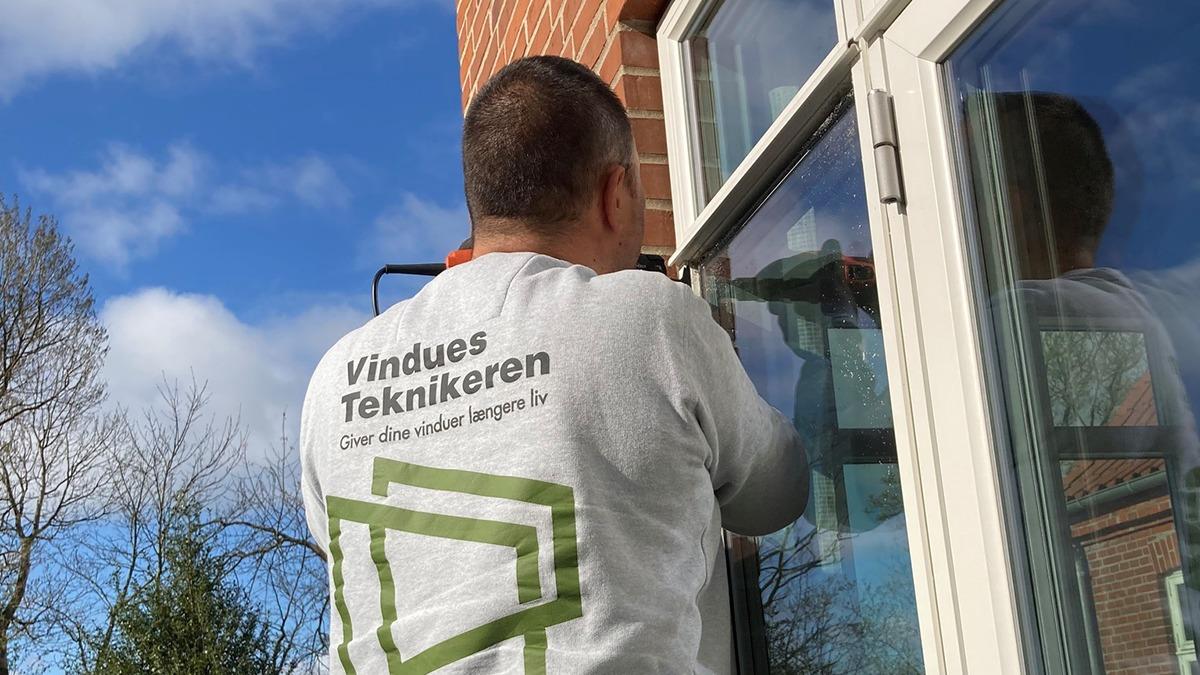 En håndværker arbejder på at reparere et vindue, iført en grå trøje med teksten "Vindues Teknikeren".