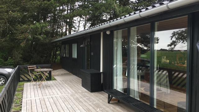 Moderne sommerhus med træterrasse, store vinduer og udsigt til grønne omgivelser.