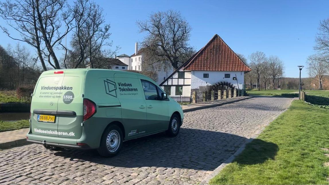En grøn varevogn parkeret på en brostensbelagt vej foran en hvid bygning med rød tag.