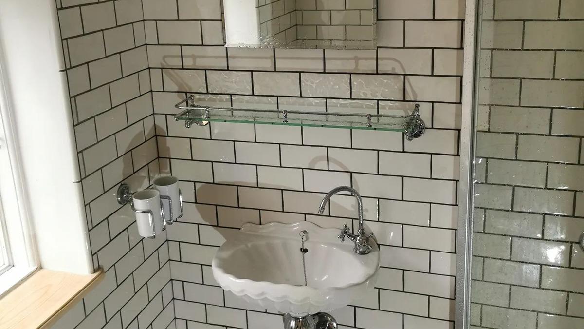 Badeværelse med hvid flisevæg, håndvask og spejl, samt en glas hylde til opbevaring.