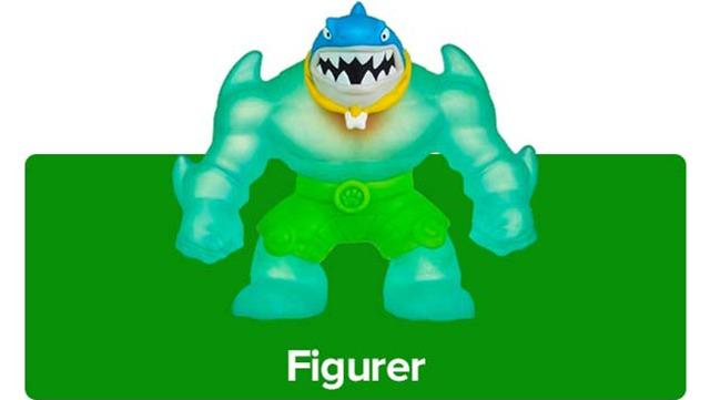 Farverig plastfigur af en haj med muskuløse arme og grønne shorts, stående på en grøn baggrund.