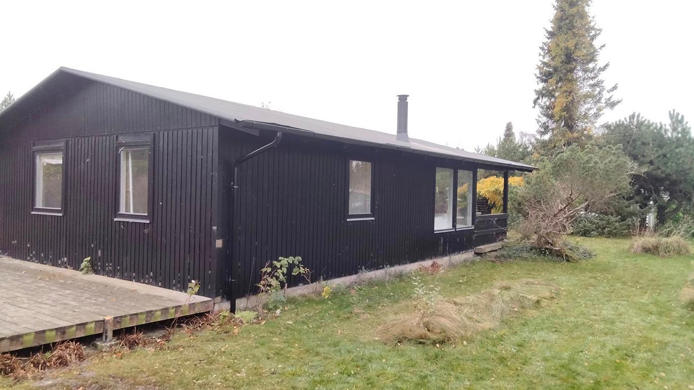 Sort træhus med terrasse og have, omgivet af grønne planter og træer.