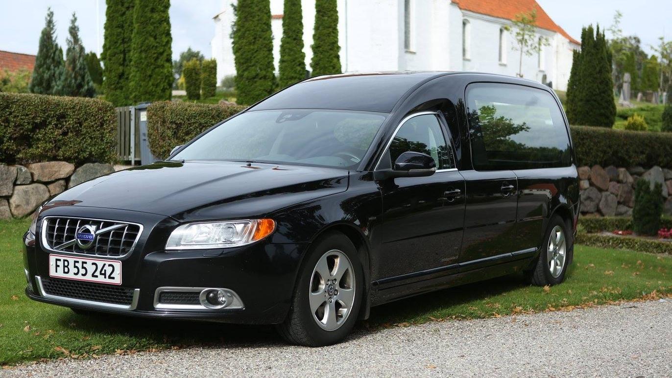 En sort Volvo stationcar parkeret ved en grusvej med grønne buske og træer i baggrunden.