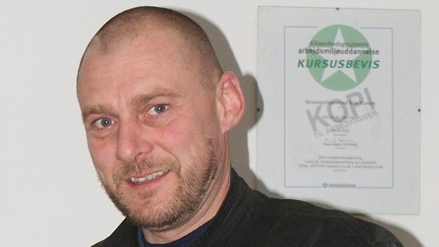Mand med kort hår smilende foran et kursusbevis hængende på væggen.