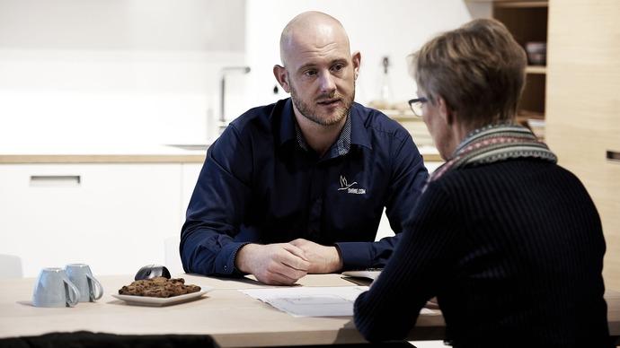 Mand i blå skjorte sidder ved bord og taler med kvinde, der har briller og stribet tørklæde, med småkager på bordet.