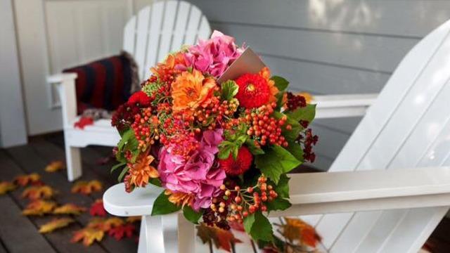 En farverig buket med pink og orange blomster ligger på en hvid stol, omgivet af efterårsblade.