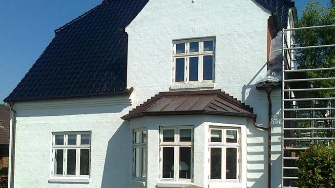 Hvidt hus med skråtag og mange vinduer, der står under solskin, med stillads ved siden af.