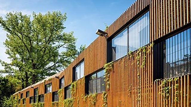 Moderne bygning med træfacade, store vinduer og klatreplanter, omgivet af grønne træer under en blå himmel.