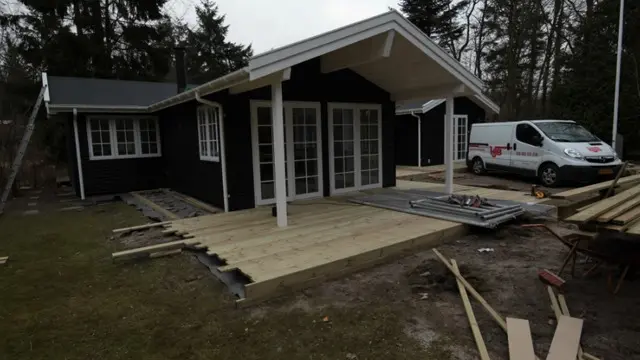 Nybygget hus med sort facade, store vinduer og træterrasse, omgivet af skov og byggeplads.