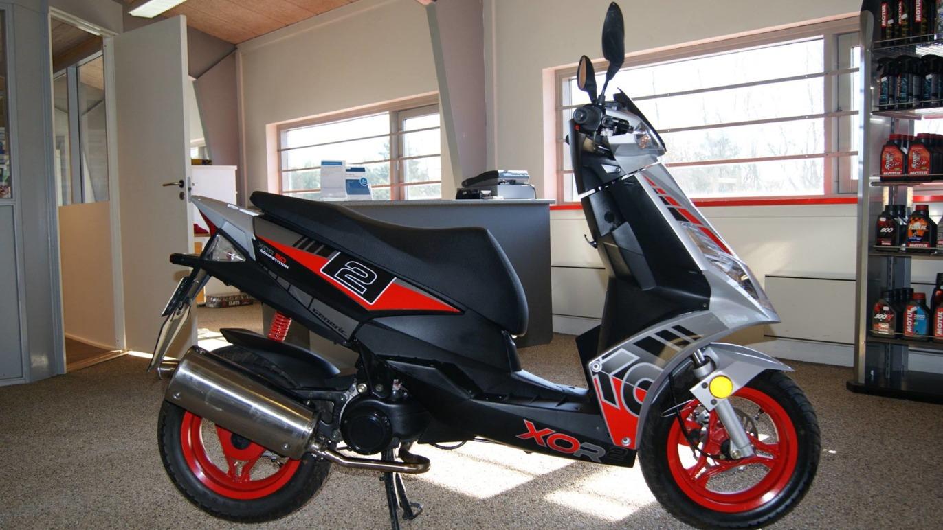 En sort og rød scooter står i et lyst rum med vinduer og hylder fyldt med bilplejeprodukter.