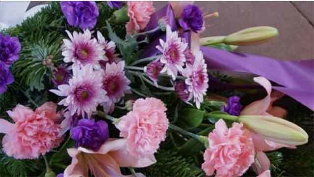 En farverig buket med lyserøde og lilla blomster, herunder nelliker og liljer, bundet med et lilla bånd.