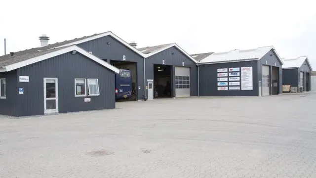 En grå bygning med flere garageporte og skilte, beliggende på en grusplads.