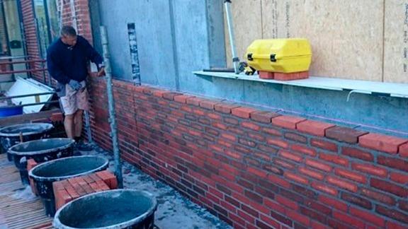 Murer arbejder med røde mursten på en byggeplads, mens han bruger en murerske og har værktøj ved siden af.